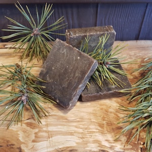 Può includere: Tre saponette di colore marrone scuro con un aspetto maculato, appoggiate su una superficie di legno. Il sapone è circondato da rami di pino con aghi verdi.