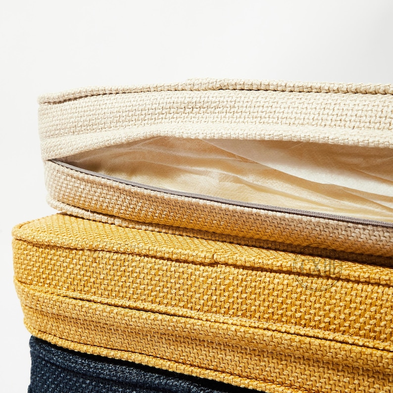 Op de afbeelding: Close-up van drie gestapelde ronde kussens in verschillende kleuren. Het bovenste kussen is beige met een ritssluiting, die een lichtgekleurd interieur onthult. Het middelste kussen is mosterdgeel en het onderste kussen is marineblauw. Alle kussens hebben een geweven textuur.