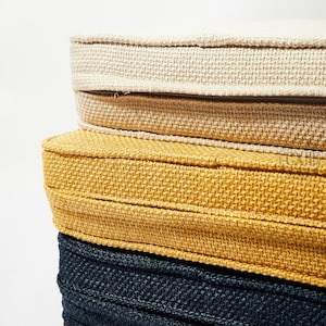 Op de afbeelding: Een stapel van vier ronde zitkussens in verschillende kleuren. Het bovenste kussen is lichtbeige, gevolgd door een bruin, een goudgeel en een diep marineblauw. Elk kussen heeft een geweven stof met textuur en een gestikte rand.