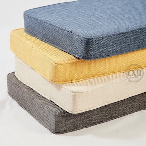 Op de afbeelding: Een stapel van vier vierkante kussens in verschillende kleuren. Het bovenste kussen is een gedempt blauw, gevolgd door geel, cr&egrave;me en donkergrijs. Elk kussen heeft een textuurstof en een eenvoudig, strak ontwerp, geschikt voor binnen of buiten.