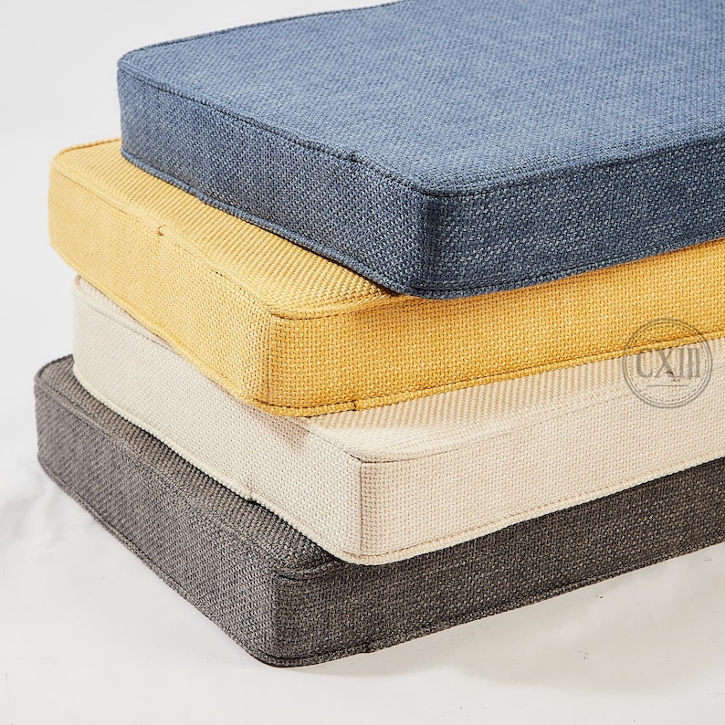 Op de afbeelding: Een stapel van vier vierkante kussens in verschillende kleuren. Het bovenste kussen is een gedempt blauw, gevolgd door geel, cr&egrave;me en donkergrijs. Elk kussen heeft een textuurstof en een eenvoudig, strak ontwerp, geschikt voor binnen of buiten.