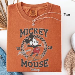 May include: An orange Comfort Colors t-shirt with a vintage Mickey Mouse graphic. The design includes the words "Mickey Mouse" and the year "1928". A silver necklace with a flower pendant is draped across the shirt. The shirt is folded neatly.