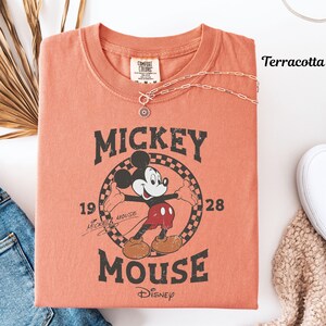 May include: A terracotta-coloured t-shirt with a vintage Mickey Mouse graphic. The design includes the text "Mickey Mouse" and the year "1928". A silver necklace with a pendant rests on the shirt. The shirt is folded, with a pair of jeans and a white shoe visible.