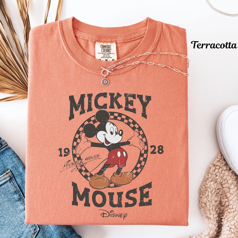 May include: A terracotta-coloured t-shirt with a vintage Mickey Mouse graphic. The design includes the text "Mickey Mouse" and the year "1928". A silver necklace with a pendant rests on the shirt. The shirt is folded, with a pair of jeans and a white shoe visible.