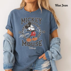 May include: Blue jean coloured t-shirt with a distressed Mickey Mouse graphic. The design includes the text "Mickey Mouse" and the year "1928". The shirt has rolled-up sleeves and a relaxed fit.