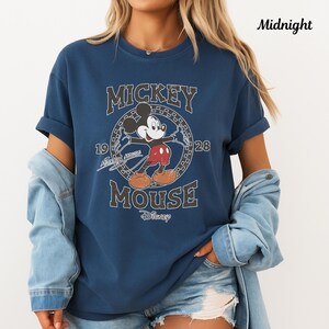 May include: A navy blue t-shirt with a vintage Mickey Mouse graphic. The design includes the words "Mickey Mouse" and the year "1928". The shirt has rolled sleeves and is worn with a denim jacket.
