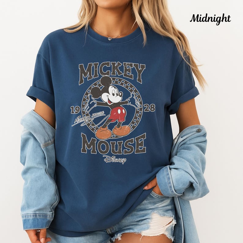 May include: A navy blue t-shirt with a vintage Mickey Mouse graphic. The design includes the words "Mickey Mouse" and the year "1928". The shirt has rolled sleeves and is worn with a denim jacket.