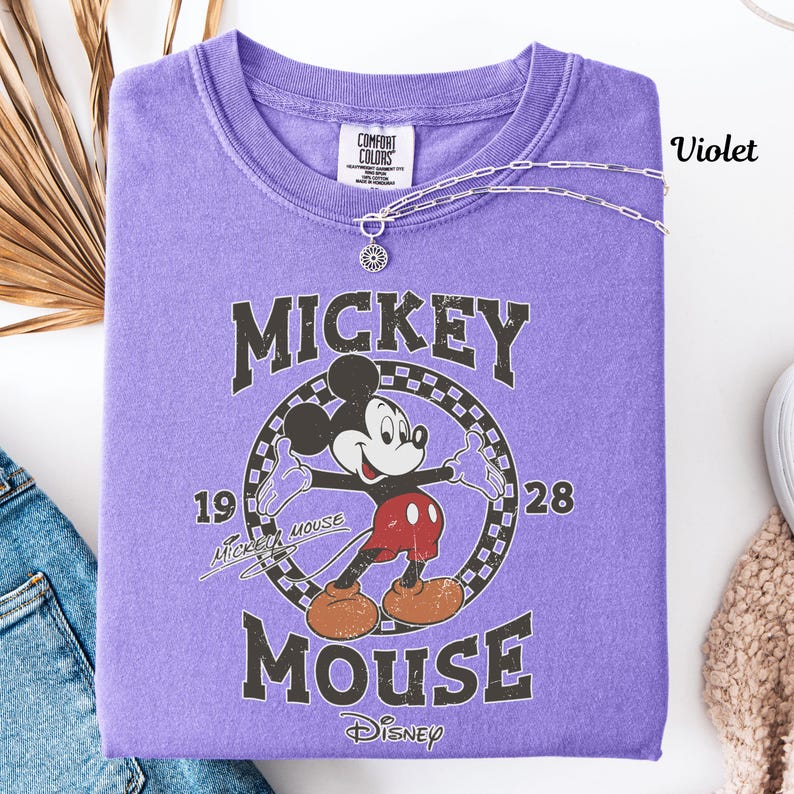 May include: A violet Comfort Colors t-shirt with a Mickey Mouse graphic. The design includes the words "Mickey Mouse" and the numbers "1928". A silver necklace with a pendant is draped across the shirt.
