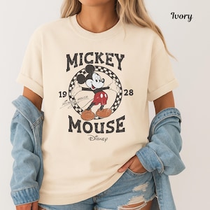 May include: Ivory-coloured t-shirt with a vintage Mickey Mouse graphic. The design includes the words "Mickey Mouse" and the year "1928" in a circular frame. The shirt is a classic, casual style.