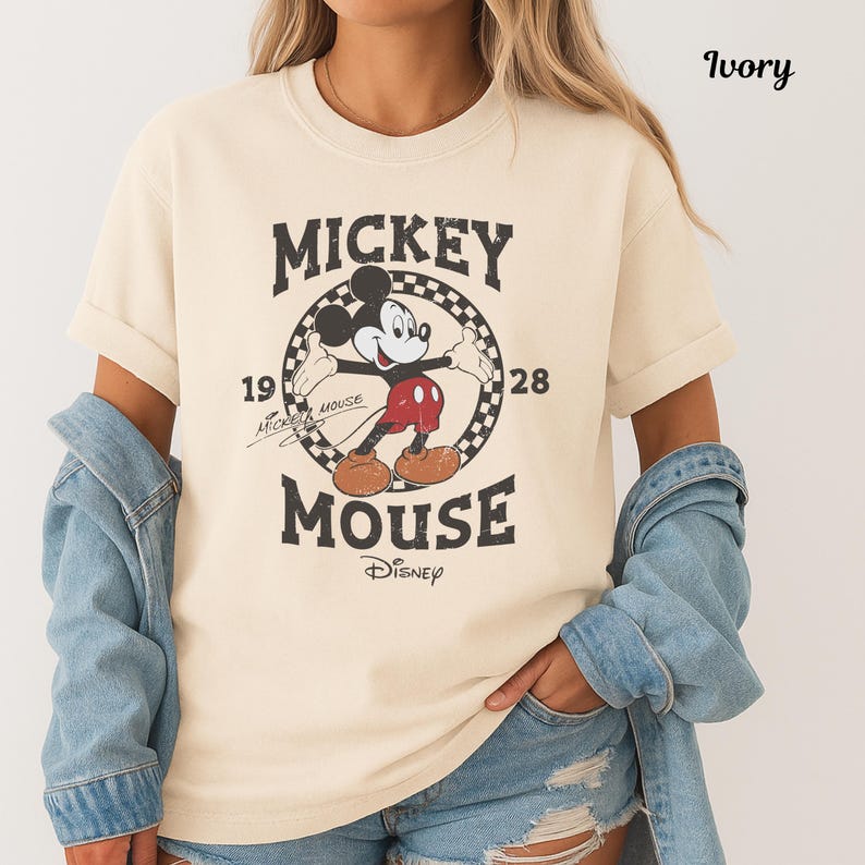 May include: Ivory-coloured t-shirt with a vintage Mickey Mouse graphic. The design includes the words "Mickey Mouse" and the year "1928" in a circular frame. The shirt is a classic, casual style.