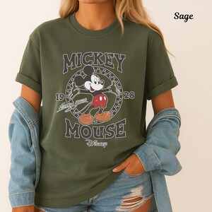 May include: Sage green t-shirt with a distressed Mickey Mouse graphic. The design includes the words "Mickey Mouse" and "1928" in a vintage style. The Disney logo is also visible on the shirt.