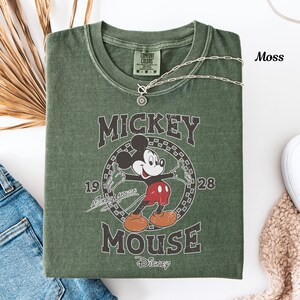 May include: A moss green t-shirt with a vintage Mickey Mouse graphic. The design includes the words "Mickey Mouse" and "Disney", along with the years "1928" and "1928". A silver necklace with a pendant rests on the shirt.