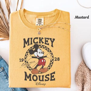 May include: Mustard-coloured t-shirt with a Mickey Mouse graphic. The design includes the words "Mickey Mouse" and "Disney" along with the year "1928". A delicate necklace rests on the shirt.