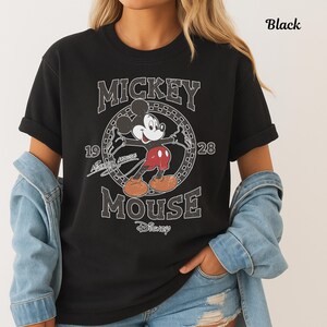 May include: Black crew neck t-shirt with a distressed Mickey Mouse graphic. The design includes the words "Mickey Mouse" and the years "1928". The Disney logo is also present. The shirt has a vintage aesthetic.