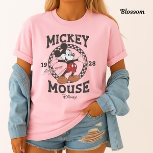 May include: A pale pink t-shirt with a Mickey Mouse graphic. The design includes the words "Mickey Mouse" and "Disney" along with the years "1928" and "19". The shirt is worn with a denim jacket and denim shorts.