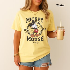 May include: A yellow t-shirt with a vintage Mickey Mouse design. The graphic includes "Mickey Mouse" and the years "1928" and "19". The shirt is worn with denim shorts. A glass of a yellow drink is held in the hand.