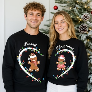 May include: Black sweatshirts with matching designs for Christmas. One sweatshirt says "Merry" with a gingerbread man wearing a hat and scarf. The other says "Christmas" with a gingerbread woman wearing a hat and dress. Both designs are framed by a white heart and string lights.