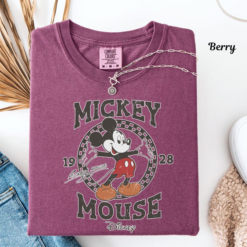 May include: Berry-coloured t-shirt with a Mickey Mouse graphic. The design includes the words "Mickey Mouse" and the year "1928". A silver necklace with a pendant is draped across the shirt. The shirt is neatly folded.