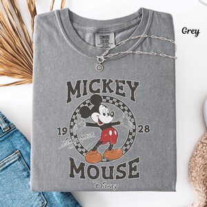 May include: A grey t-shirt with a vintage Mickey Mouse design. The graphic includes "Mickey Mouse" and the years "1928" and "19". A silver necklace with a pendant is draped over the shirt.