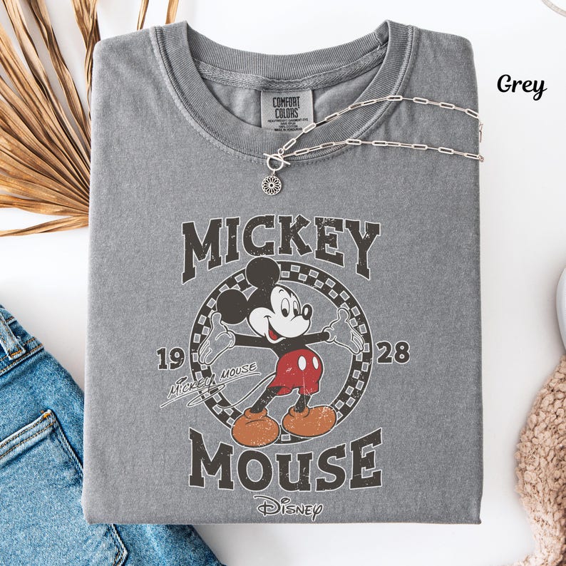May include: A grey t-shirt with a vintage Mickey Mouse design. The graphic includes "Mickey Mouse" and the years "1928" and "19". A silver necklace with a pendant is draped over the shirt.