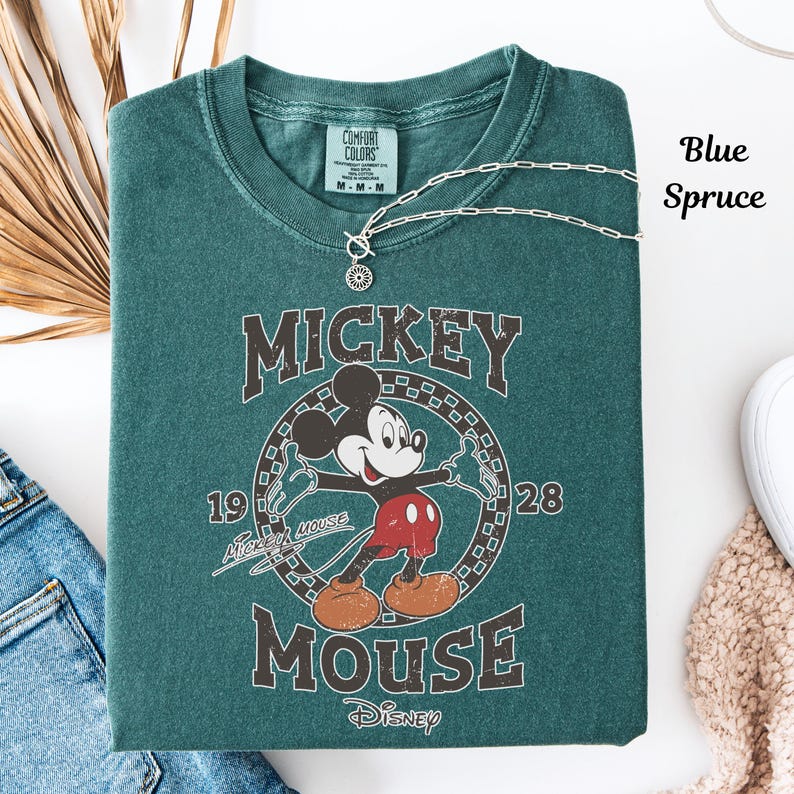 May include: Blue spruce-coloured t-shirt with a vintage Mickey Mouse graphic. The design includes Mickey Mouse, the words "Mickey Mouse" and "Disney", and the years "1928" and "19". A silver necklace with a pendant is draped over the shirt.