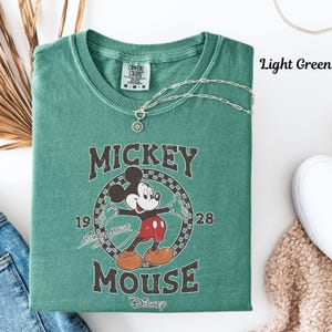 May include: Light green t-shirt with a vintage Mickey Mouse graphic. The design includes Mickey Mouse, the words "Mickey Mouse" and the years "1928" and "1919". A silver necklace with a pendant is draped over the shirt. The text "Light Green" is visible.