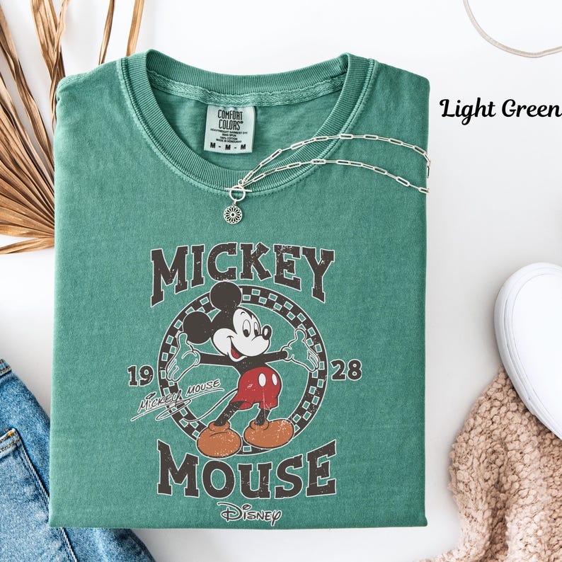 May include: Light green t-shirt with a vintage Mickey Mouse graphic. The design includes Mickey Mouse, the words "Mickey Mouse" and the years "1928" and "1919". A silver necklace with a pendant is draped over the shirt. The text "Light Green" is visible.