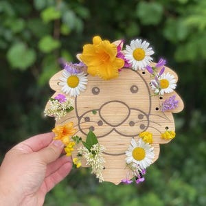 May include: A wooden lion-shaped craft with a painted face, adorned with colorful flowers. The lion's mane is decorated with yellow, white, and purple flowers. The craft is held against a blurred green background.