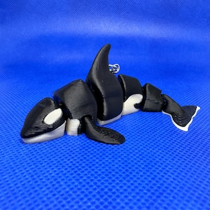 Può includere: Un portachiavi a forma di orca stampato in 3D in bianco e nero. L'orca è in posizione di nuoto con la pinna dorsale sollevata. Il portachiavi è in plastica e ha un anello di metallo argentato.