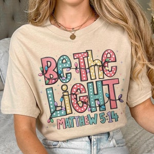 Puede incluir: Camiseta color arena con el texto "Be the Light" en letras coloridas y luces de cadena. El texto "Matthew 5:14" está debajo. La camiseta es de estilo casual.