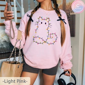 Adorable light pink sweatshirt featuring a minimalist cat outline wrapped in colorful Christmas lights. Perfect for adding festive cheer and pastel charm to holiday gatherings or winter outings.