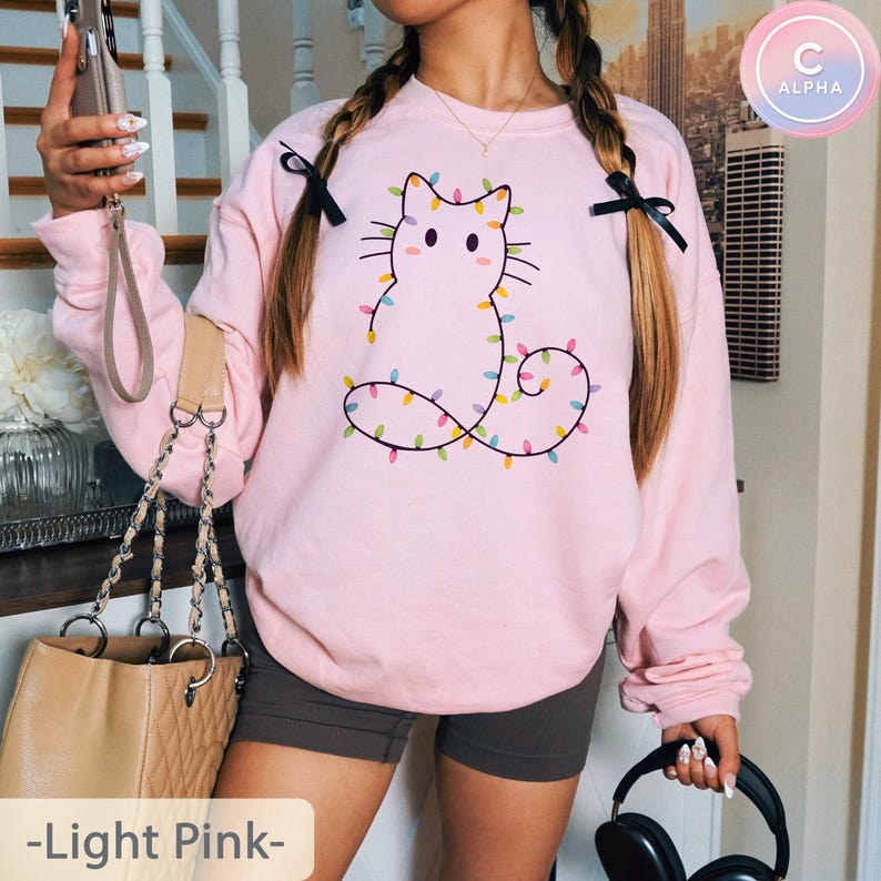 Adorable light pink sweatshirt featuring a minimalist cat outline wrapped in colorful Christmas lights. Perfect for adding festive cheer and pastel charm to holiday gatherings or winter outings.
