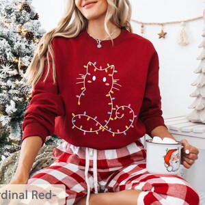 Charming cardinal red sweatshirt featuring a festive cat outline with Christmas lights. A cute and cheerful addition to holiday parties and winter celebrations.