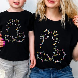Matching black T-shirts for adults and kids featuring a festive cat outline with colorful Christmas lights. Perfect for family photos and holiday memories.