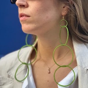Puede incluir: Un par de aretes largos de aro de cuentas verdes. Los aretes están hechos de múltiples círculos de cuentas, con el círculo más grande en la parte inferior. Los aretes cuelgan de los lóbulos de las orejas de una persona que lleva una camisa blanca y una chaqueta marrón.