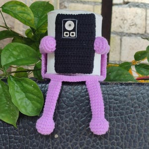 May include: A handmade, crocheted plush toy in the shape of a smartphone. The toy features a black screen with white details, a white border, and purple arms and legs. The legs are dangling, and the toy is sitting on a textured surface.