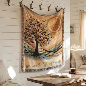 May include: A tapestry with a tree and sun design, in shades of brown, orange, and blue, hangs on a white wall. The tapestry has a fringed edge and the text "you are the light of my life" at the bottom. Decorative wall hanging.