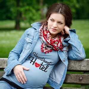 May include: A pregnant person wearing a light blue t-shirt with the text "Heartbeat Hotel" and a red heart with a heartbeat graphic. The person is wearing a red and white plaid scarf with red pom-poms and a blue denim jacket.