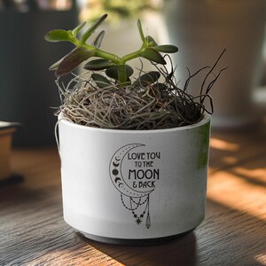 May include: White ceramic planter with a green and white speckled glaze. The planter has a black graphic of a crescent moon with stars and a quote that reads "Love you to the moon & back".