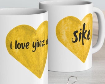 May include: Two white ceramic mugs with black text and yellow heart designs. One mug says "i love yinz d" and the other says "sike". The mugs have a glossy finish and a curved handle.