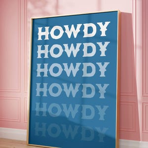 May include: A framed art print with the word "HOWDY" repeated six times in white, bold, serif font on a blue background. The frame is gold, and the artwork leans against a pink wall with a wooden floor.