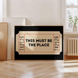 Puede incluir: Impresión artística enmarcada con un diseño de boleto vintage. El boleto tiene el texto "THIS MUST BE THE PLACE" en letras negras en negrita. El borde del boleto es rojo con el número "742126" impreso en cada lado. El fondo es negro.