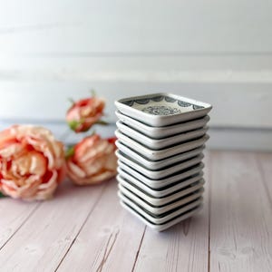 May include: A stack of square, white ceramic dishes with a black and blue floral design on the top surface. The dishes are stacked on top of each other. Soft pink roses are in the background.
