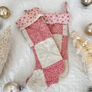 May include: Two handmade Christmas stockings. The stockings are red with a small white floral pattern and cream-colored patchwork. The cuffs are pink and white striped with red polka dots. The stockings are decorated with vintage ornaments and small white Christmas trees.
