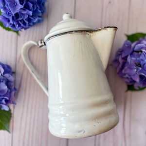 May include: White enamel coffee pot with a worn, distressed finish. The pot has a rounded body and a spout on one side. The handle is attached to the side of the pot.