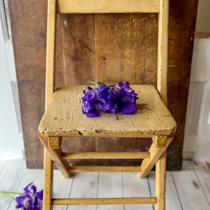 May include: A small, antique wooden chair with a distressed, cream-colored finish. A cluster of vibrant purple flowers rests on the seat. The chair is set against a rustic wooden backdrop, creating a vintage aesthetic.