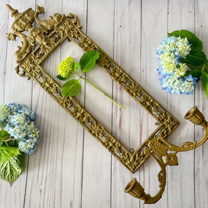 May include: Ornate gold metal wall sconce with two candle holders and a decorative frame. The frame is decorated with floral motifs and has a delicate floral arrangement inside.