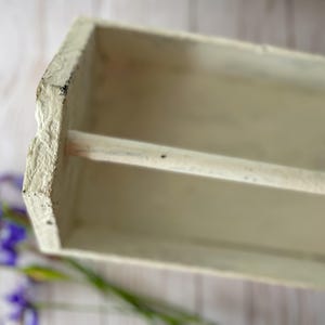May include: A rectangular, off-white wooden tray with a distressed finish. The tray has a single divider and a rough, textured edge. The background includes purple flowers and a light-colored wooden surface.