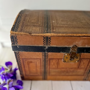 May include: A vintage brown trunk with a decorative pattern and black trim. The trunk features a metal clasp and hinges, suggesting it is an antique storage item. The design includes rectangular and X-shaped details, with a dark border.