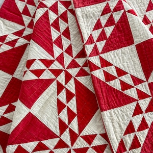 May include: A folded quilt with a striking geometric pattern. The design features alternating red and white triangles arranged in a radial pattern. The quilt's texture is visible, suggesting a handmade or vintage quality. The overall aesthetic is a blend of traditional and modern design.